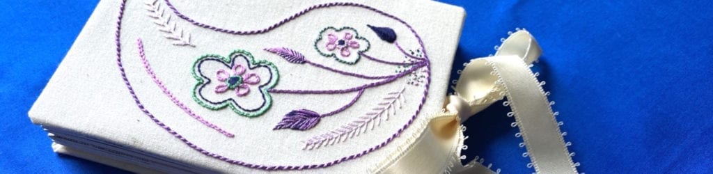 An example of what a student could learn during an embroidery course. It features a rectangular canvas on a blue background and the canvas had a purple Paisley pear with purple and turquoise flowers.