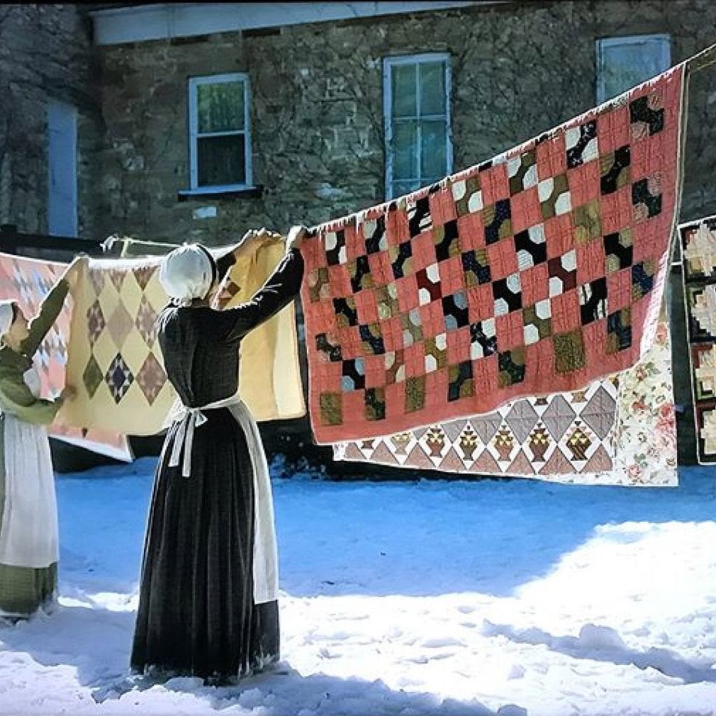 A washing line full of brightly patterned patchwork quilts. Two women hang them up while snow covered the ground.